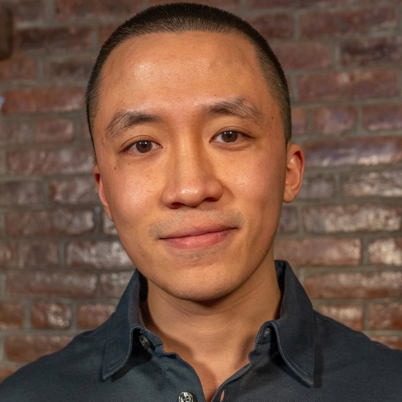 A man with a buzz cut and a dark collared shirt smiles slightly in front of a brick wall.
