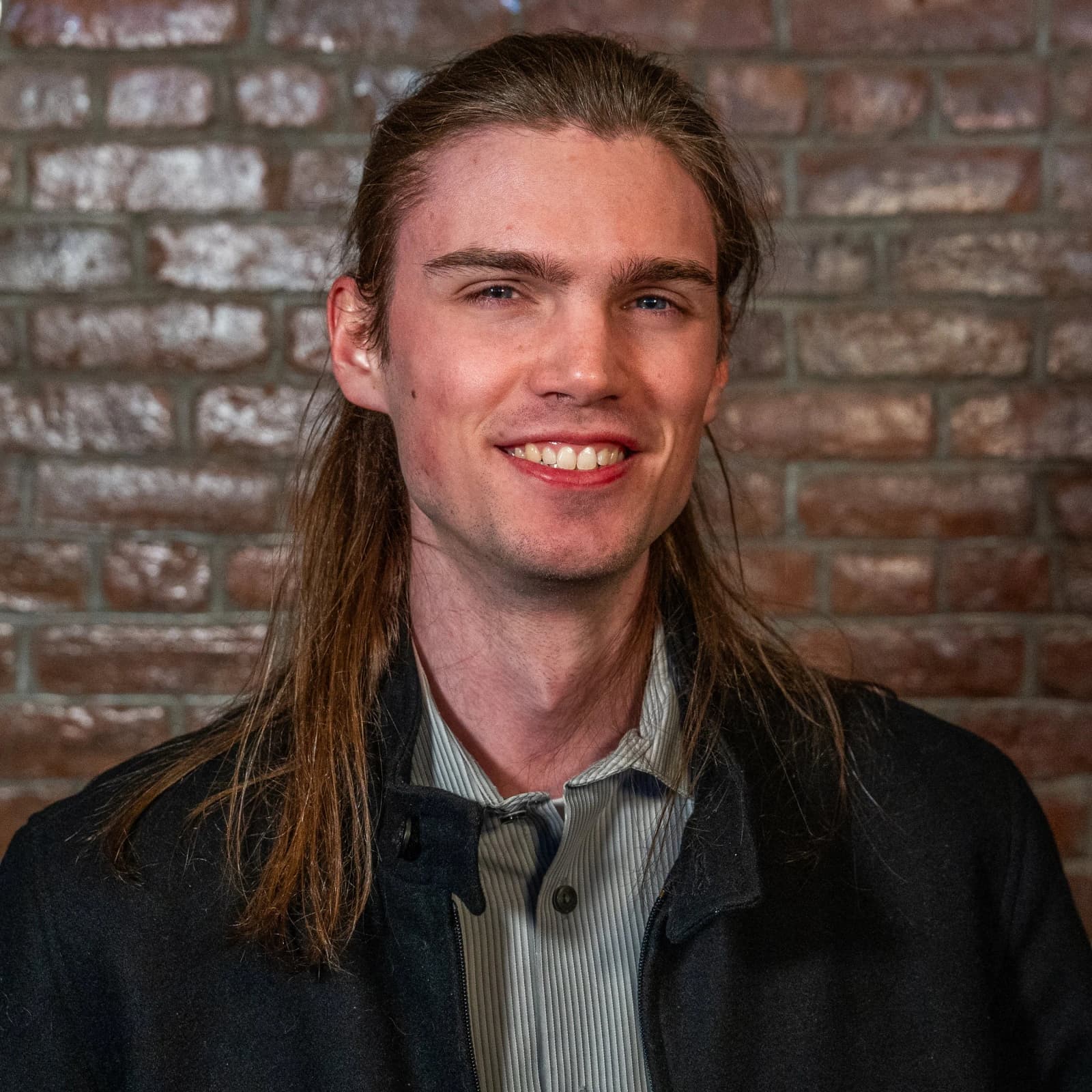 A smiling man with long hair, in a dark jacket and striped shirt, stands in front of a brick wall.