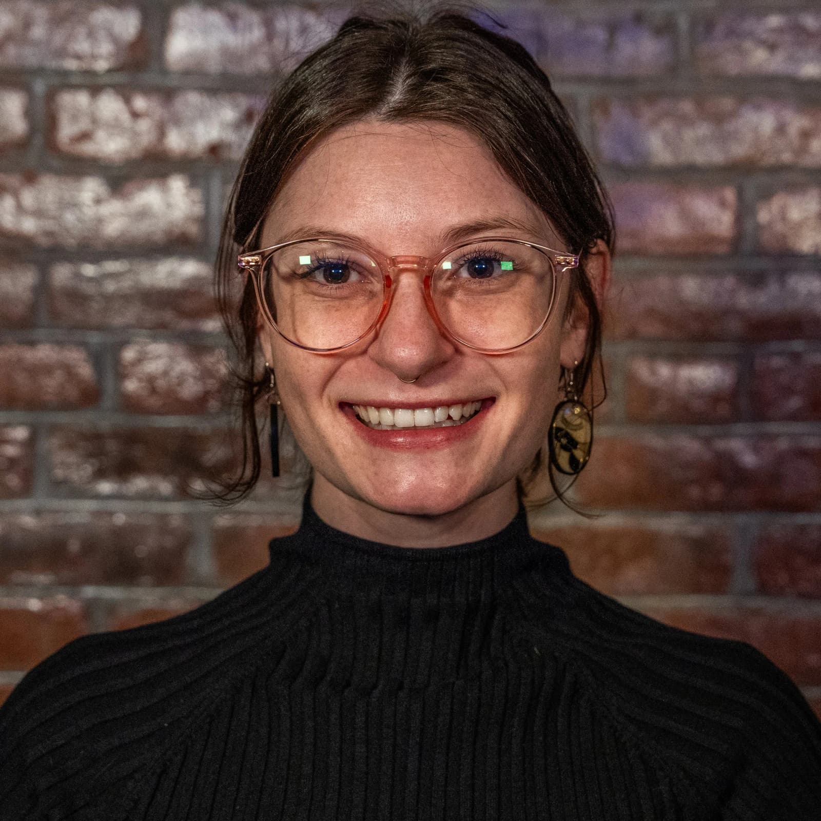 A smiling woman in glasses and a black ribbed turtleneck stands in front of a brick wall.