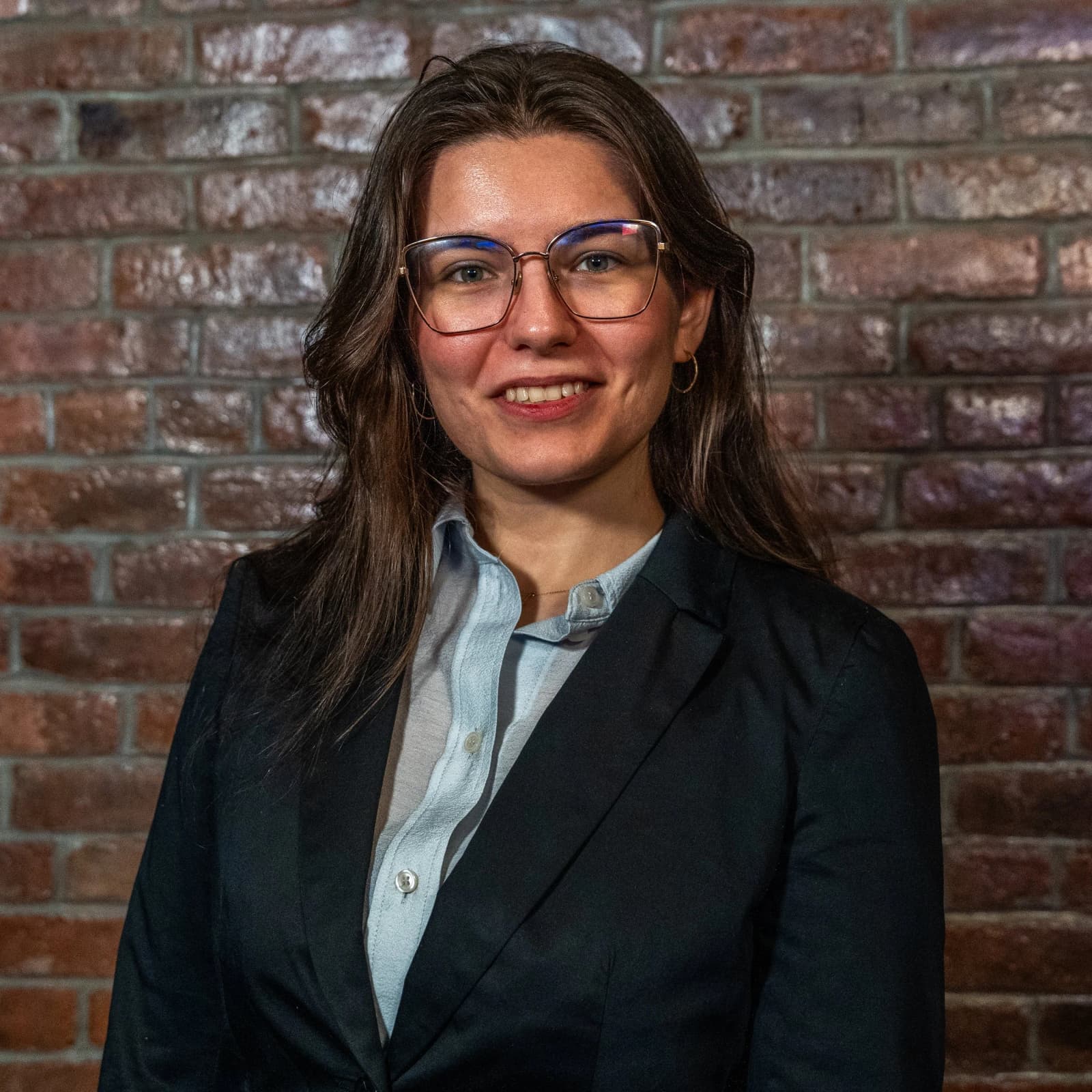 A smiling woman in glasses, a black blazer and a light blue shirt stands in front of a brick wall.