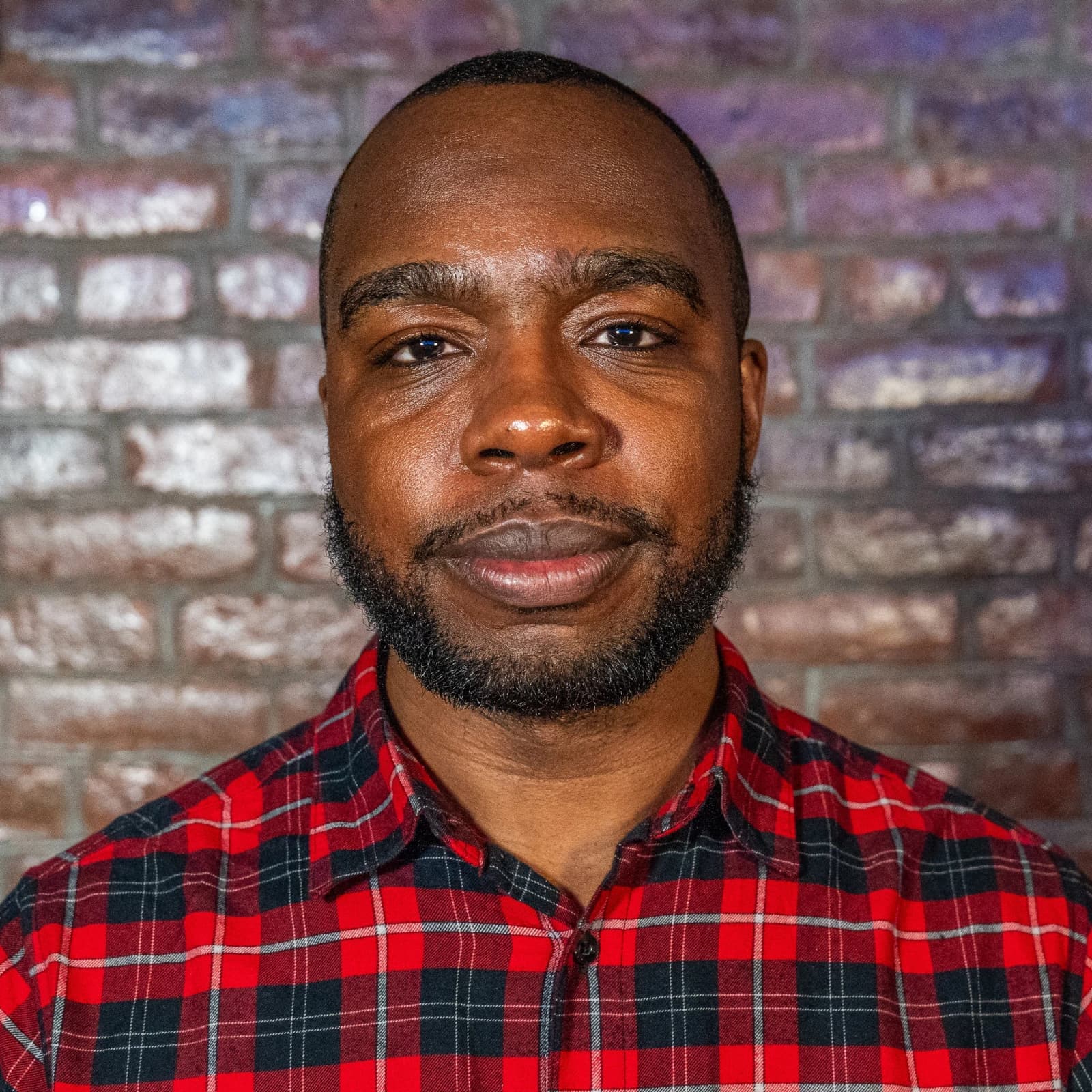 A bearded man in a red plaid shirt looks into the camera in front of a brick wall.