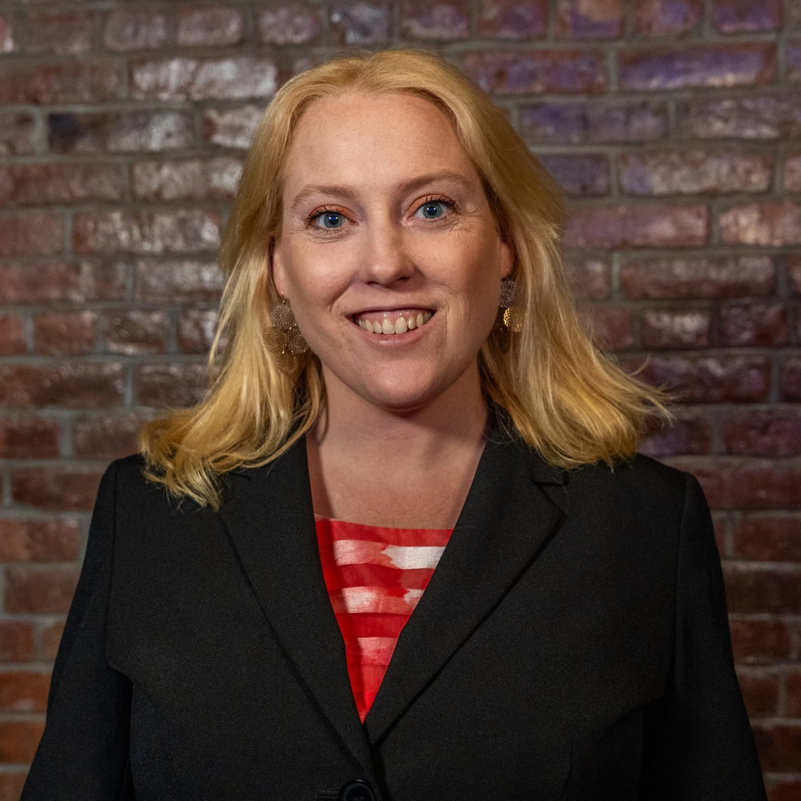 A smiling blond woman in a black blazer over a red striped top stands in front of a brick wall.