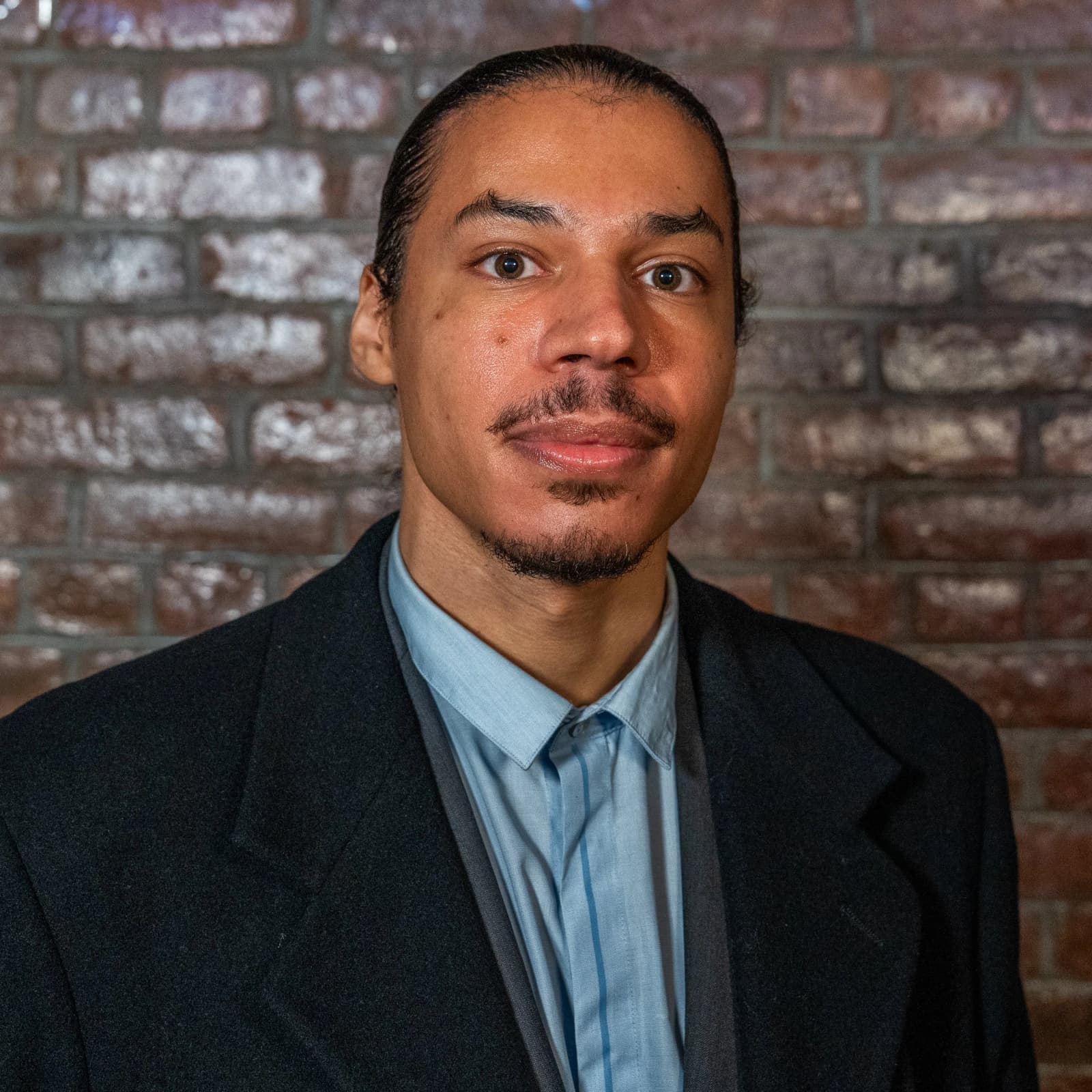 A man with slicked-back hair and a goatee, in a dark blazer and light blue shirt, stands in front of a brick wall.