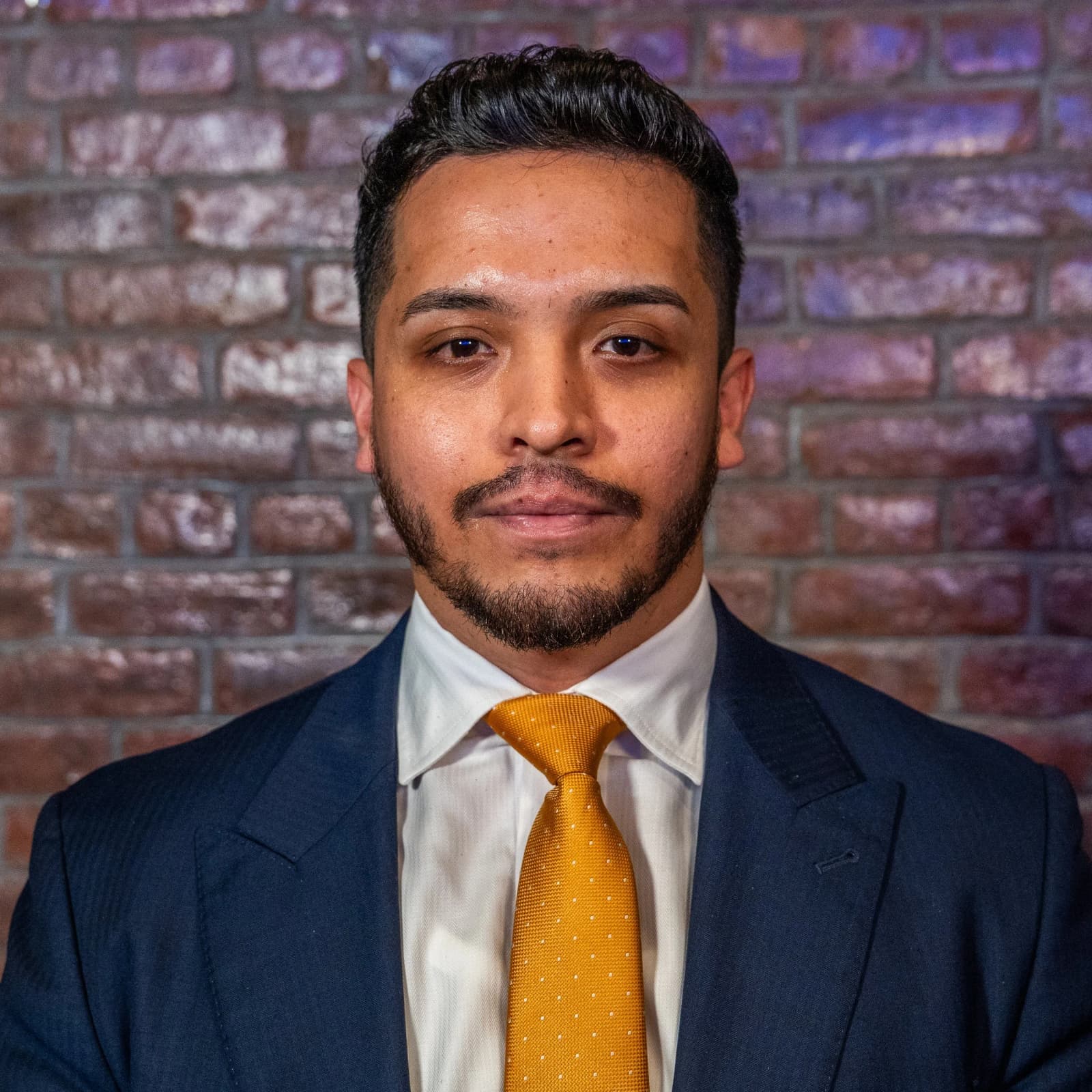 A bearded man in a navy suit, white shirt and gold tie stands in front of a brick wall.