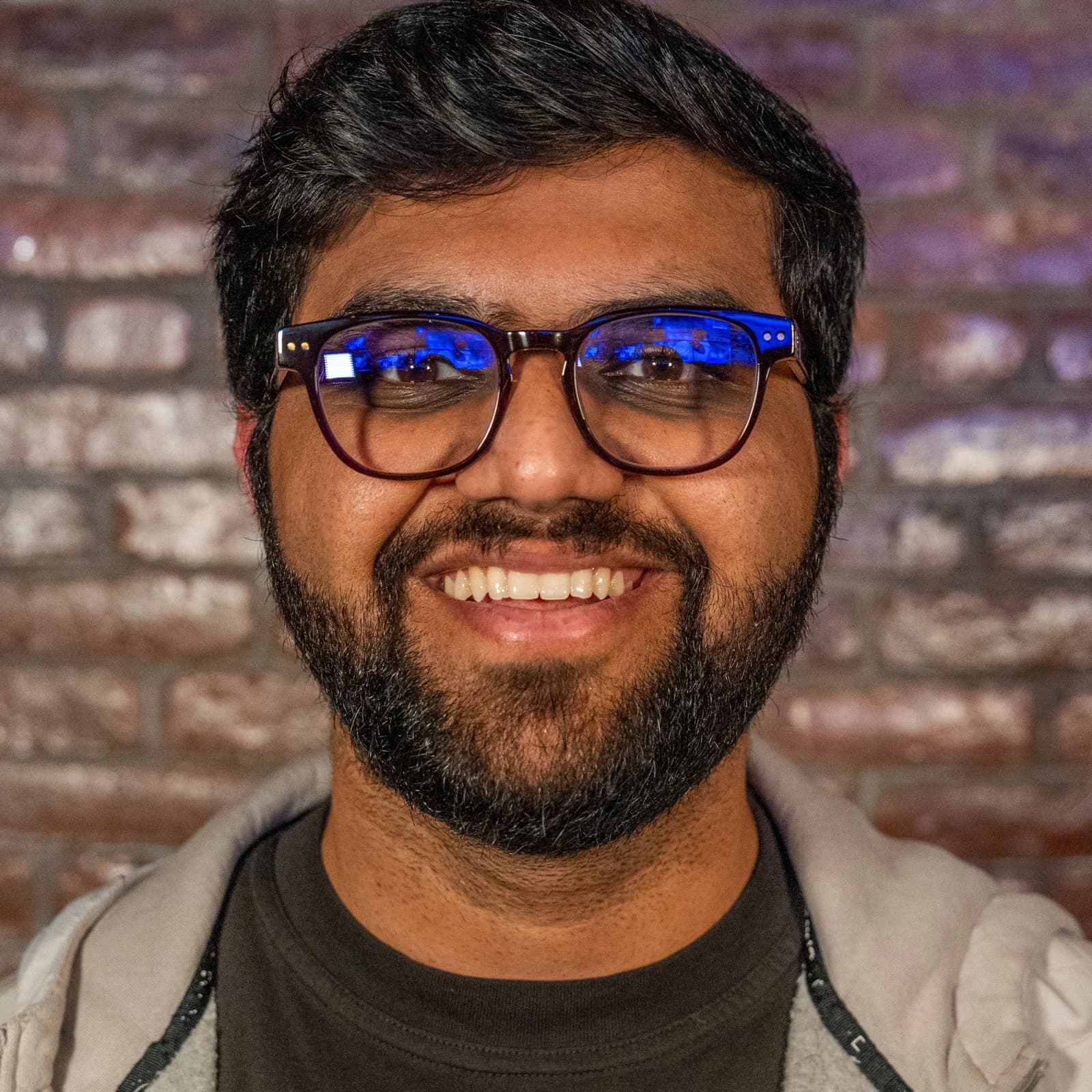 A smiling bearded man in glasses and a light gray hoodie stands in front of a brick wall.