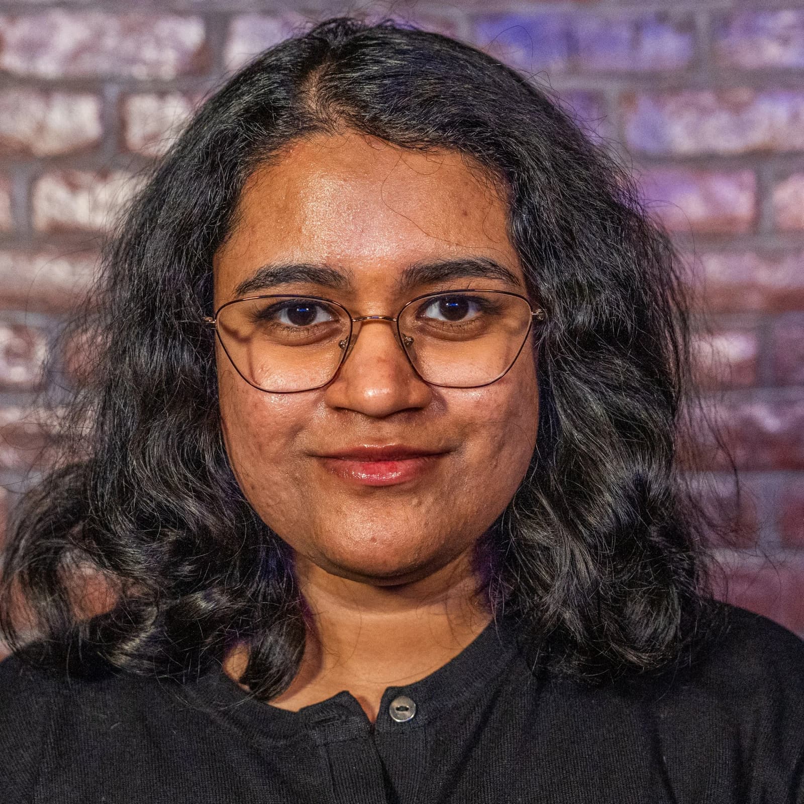 A woman with shoulder-length wavy hair and glasses, in a black top, smiles softly in front of a brick wall.