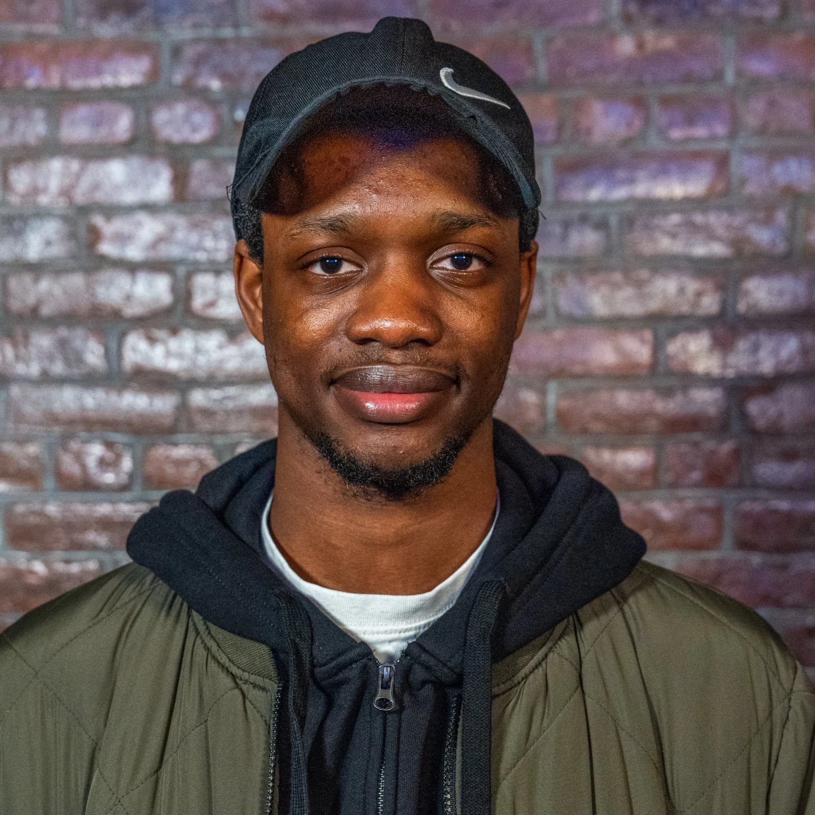 A man in a black cap, black hoodie and olive jacket smiles slightly in front of a brick wall.