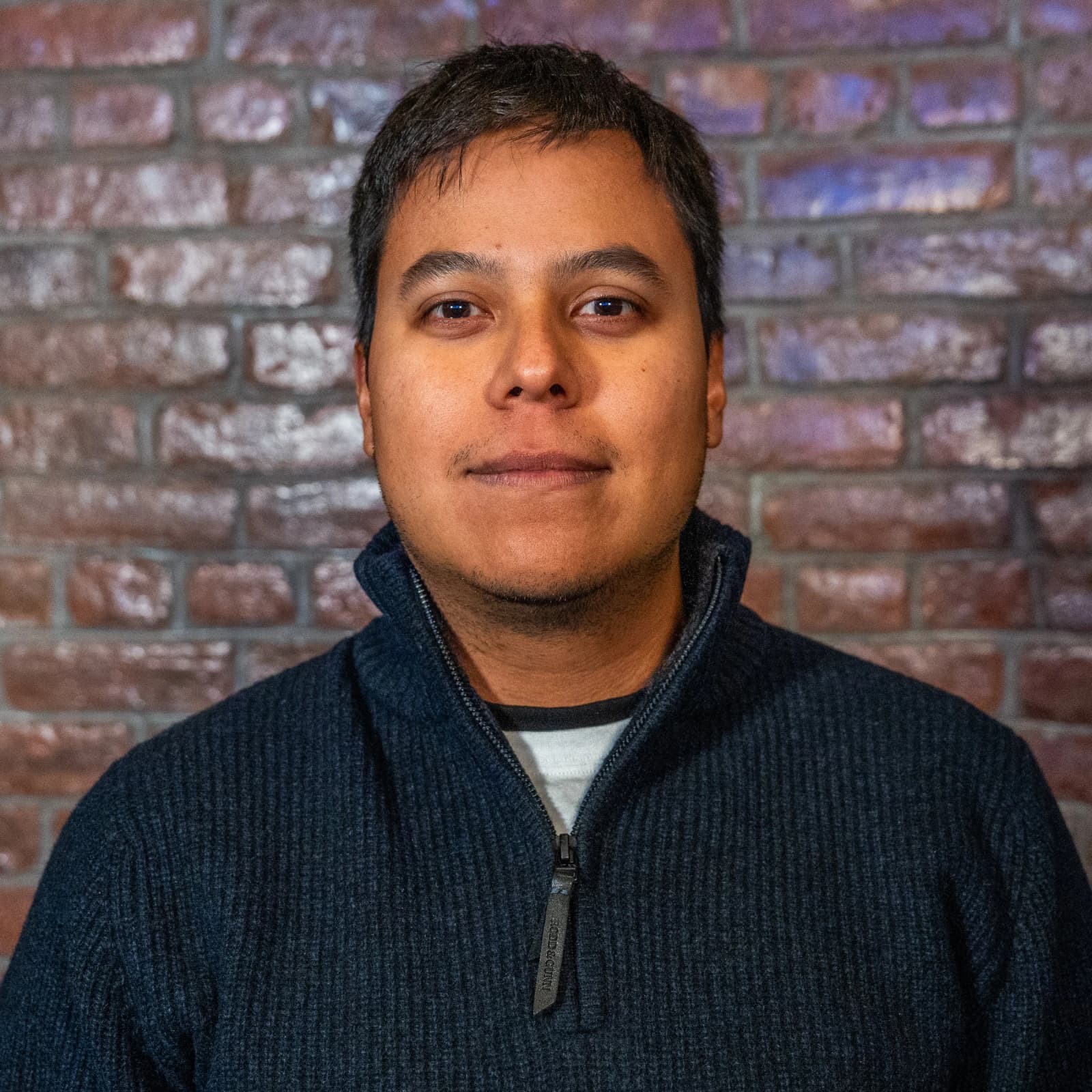 A man with short dark hair in a navy quarter-zip sweater smiles slightly in front of a brick wall.