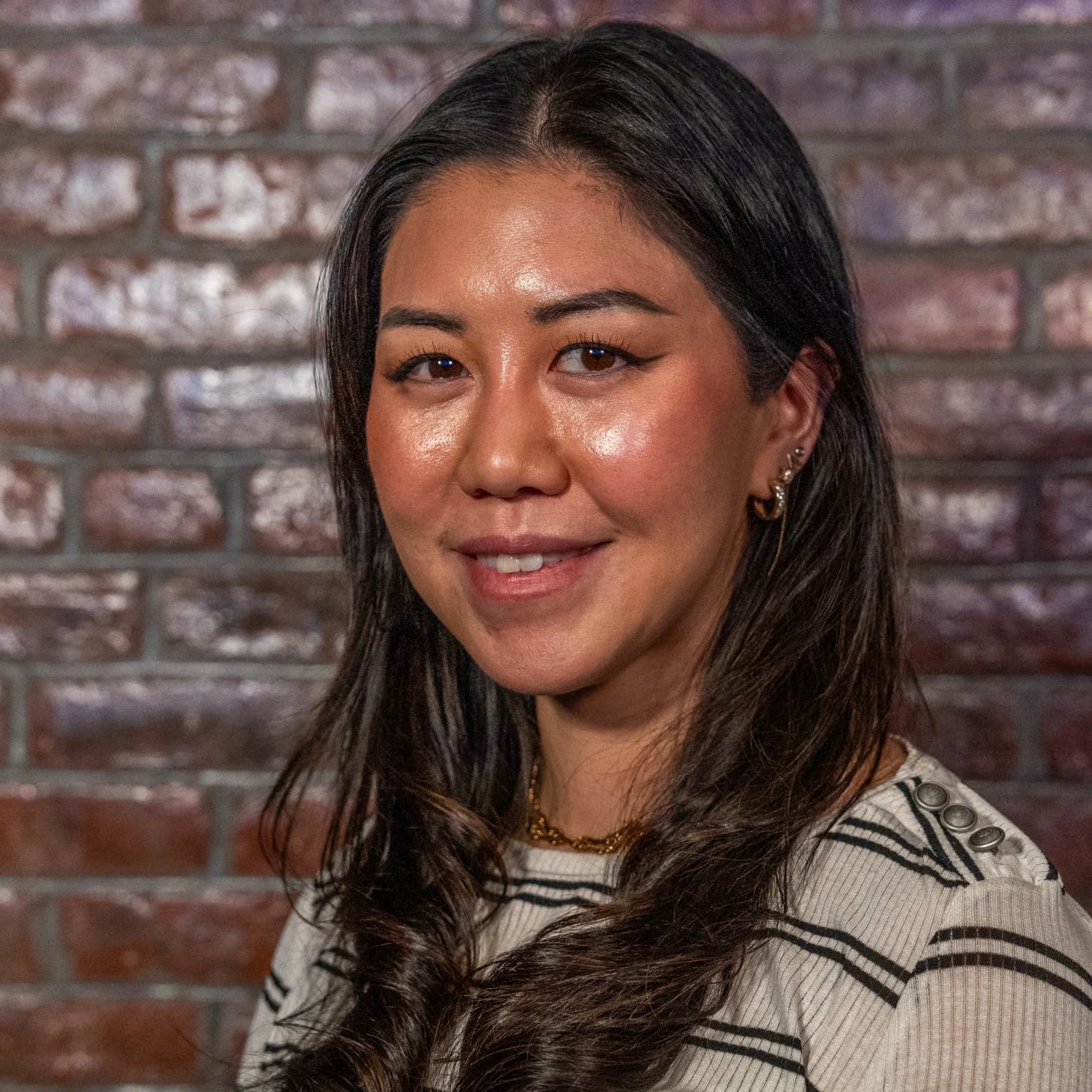 A smiling woman with long dark hair in a cream striped sweater stands in front of a brick wall.
