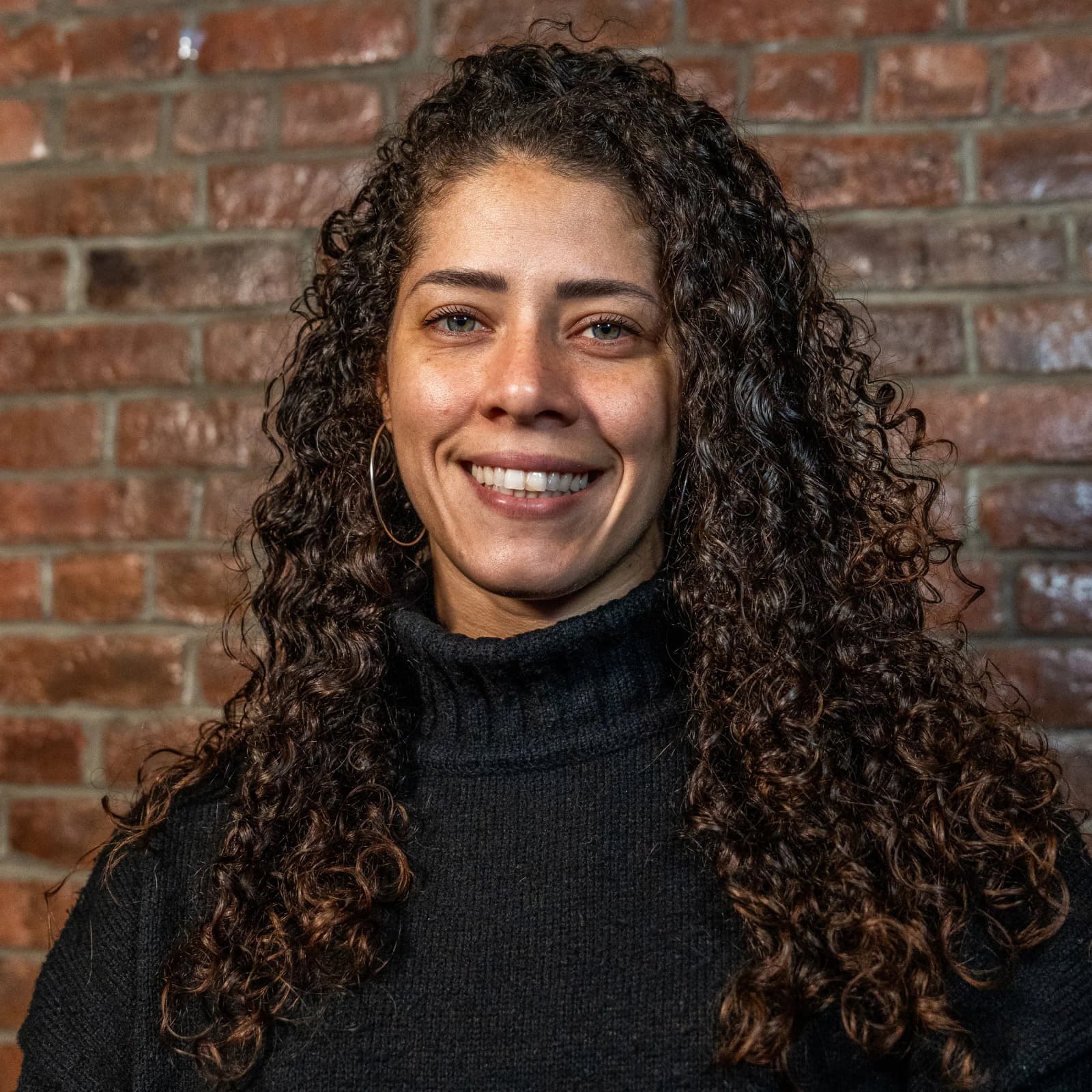 A smiling woman with long curly hair and hoop earrings, in a black turtleneck, stands in front of a brick wall.