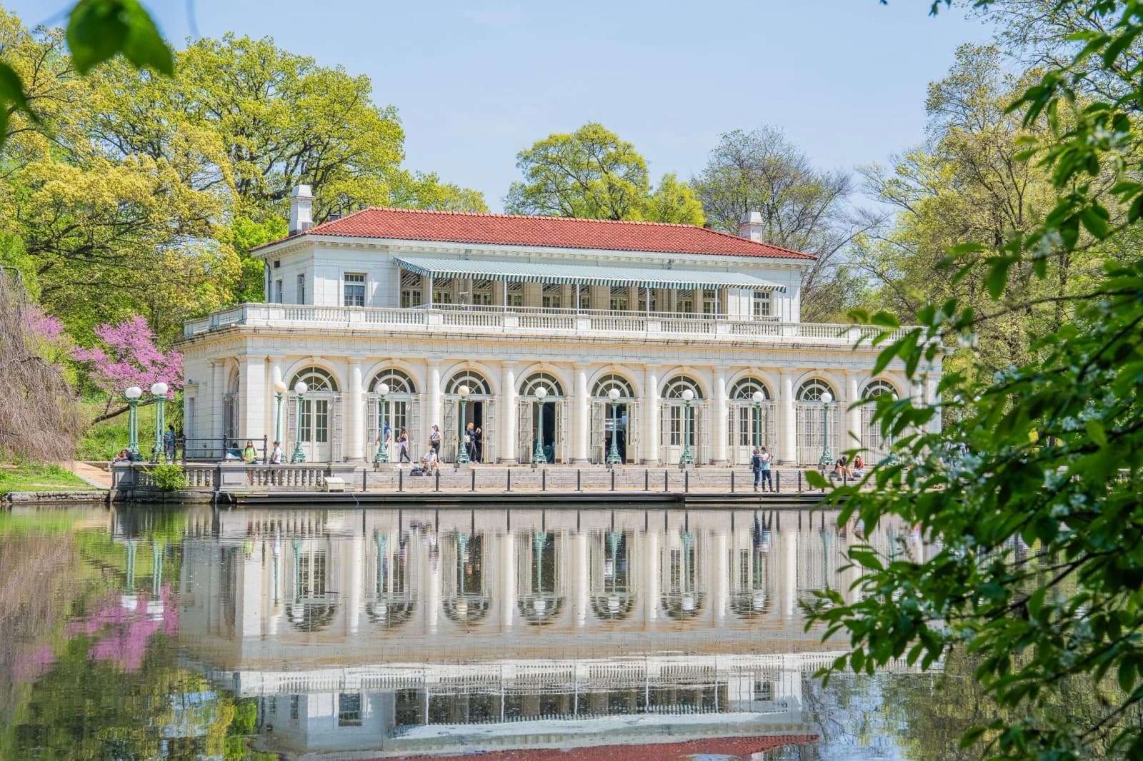 A white Beaux-Arts boathouse with arched windows and a red tile roof is mirrored in a still lake, framed by spring trees.
