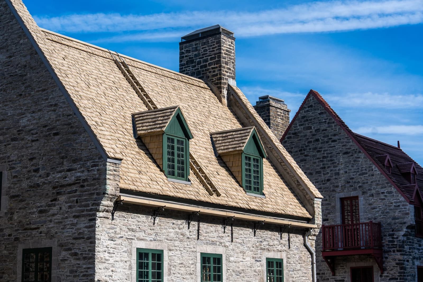 Two green-trimmed dormer windows sit on the shingled roof of a stone cottage under a blue sky.