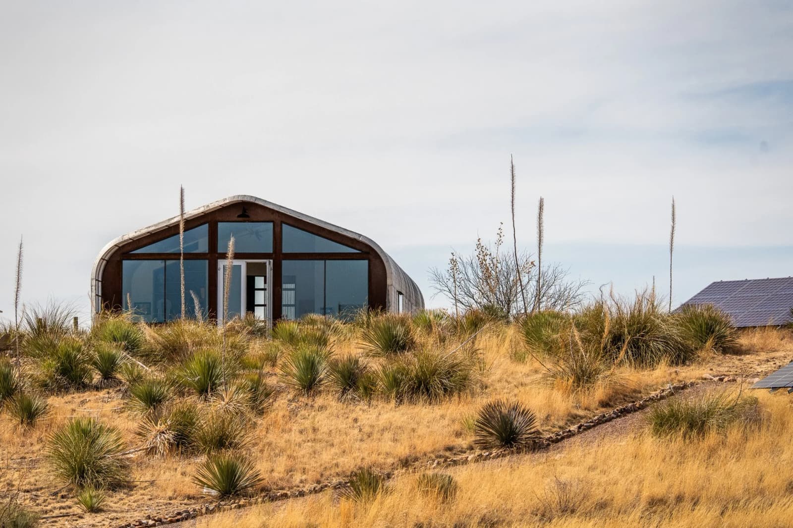 A glass-fronted, arched steel building sits on a hill of yucca and dry grass in the Arizona desert.