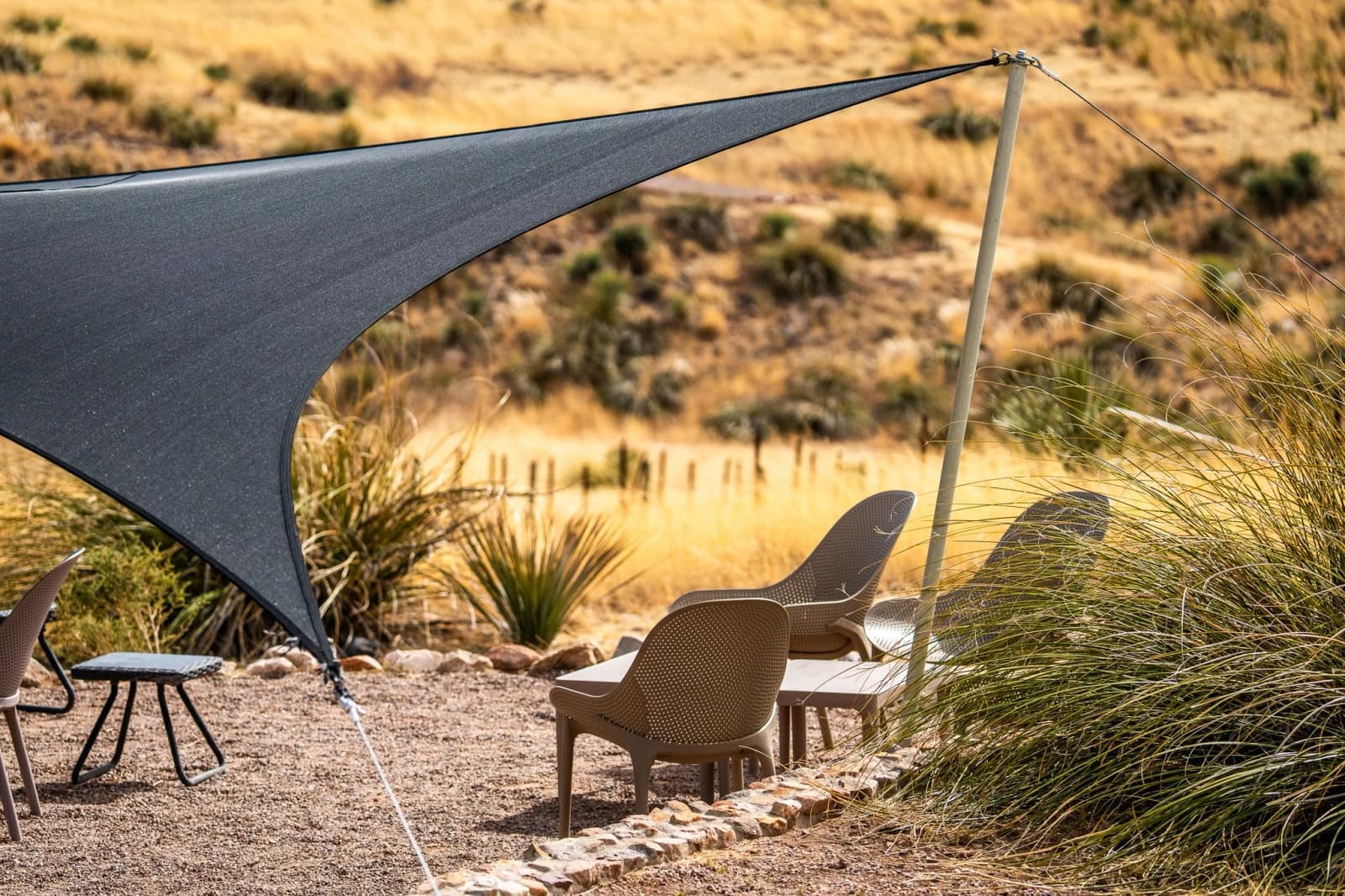 Beige wicker-style chairs sit on gravel under a stretched gray shade sail on an Arizona desert patio.