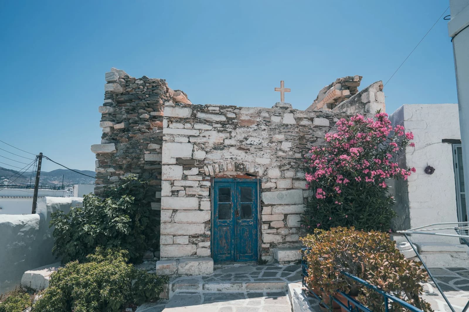 A small stone chapel with a cross on its roof and a weathered blue door stands beside pink oleander on the island of Paros in Greece.