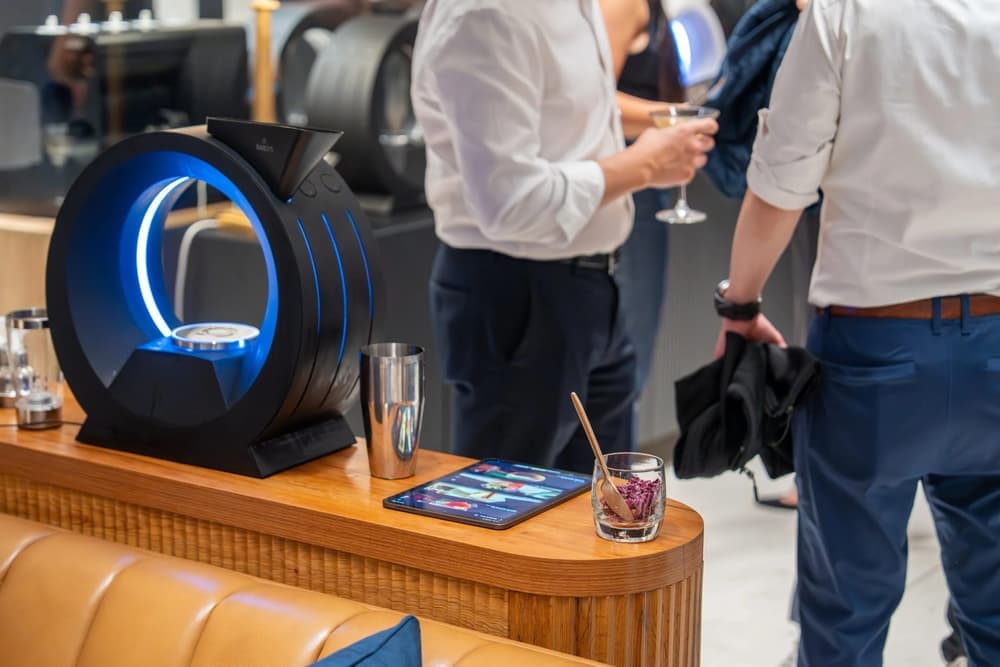 A black Barsys 360 cocktail maker glows blue on a wooden bar beside a tablet and a glass of rose petals as guests mingle at a private event.