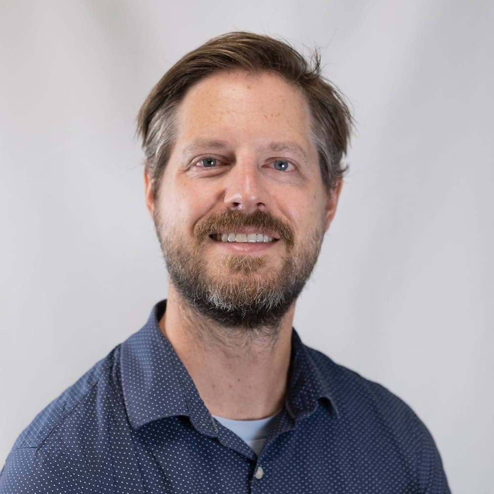 Ben Garves, smiling, with a beard and blue eyes, wearing a navy shirt with small white dots over a light blue T-shirt, in front of a plain light gray backdrop.